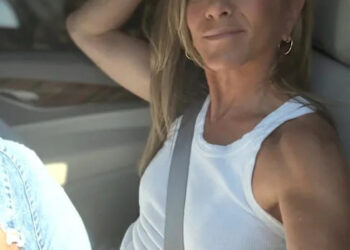 A woman with long blonde hair, wearing a white sleeveless top and blue jeans, sits in the driver's seat of a car, smiling at the camera with her arm resting on the seat back.