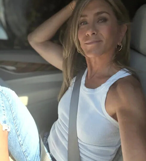 A woman with long blonde hair, wearing a white sleeveless top and blue jeans, sits in the driver's seat of a car, smiling at the camera with her arm resting on the seat back.