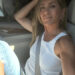 A woman with long blonde hair, wearing a white sleeveless top and blue jeans, sits in the driver's seat of a car, smiling at the camera with her arm resting on the seat back.