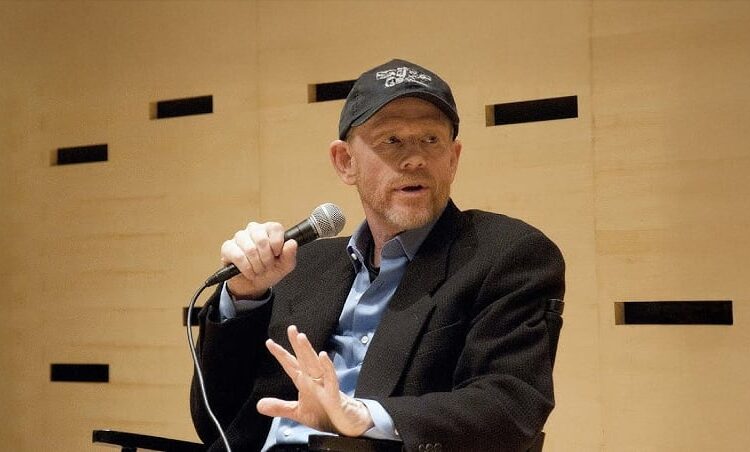 A man wearing a black baseball cap, blazer, and blue shirt holds a microphone and gestures with his hand while seated against a light wooden wall with small black rectangular accents.