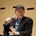 A man wearing a black baseball cap, blazer, and blue shirt holds a microphone and gestures with his hand while seated against a light wooden wall with small black rectangular accents.