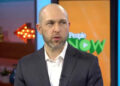 A man in a dark suit and white shirt speaks in a studio with colorful set decor. A screen behind him displays the "People NOW" logo, and a lit arrow sign is visible in the background.
