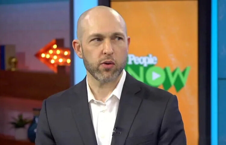 A man in a dark suit and white shirt speaks in a studio with colorful set decor. A screen behind him displays the "People NOW" logo, and a lit arrow sign is visible in the background.