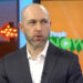 A man in a dark suit and white shirt speaks in a studio with colorful set decor. A screen behind him displays the "People NOW" logo, and a lit arrow sign is visible in the background.