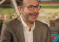 A man with glasses and a beard, wearing a brown checked blazer and white shirt, smiles while speaking. The background is colorful and blurred, suggesting a casual or interview setting.