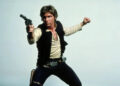 A man in a black vest, beige shirt, and dark pants strikes an action pose, aiming a blaster pistol with one arm extended and the other bent. He has brown hair and wears a holster on his hip, standing against a plain background.