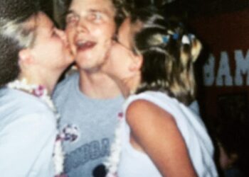 A young man wearing a gray t-shirt smiles widely as two young women, each wearing leis, kiss him on the cheeks. The background is slightly blurred with indistinct signs and decorations.