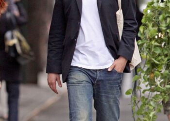 A man wearing a black fedora, sunglasses, black blazer, white t-shirt, blue jeans, and red sneakers walks on a city sidewalk with one hand in his pocket and a tote bag over his shoulder.