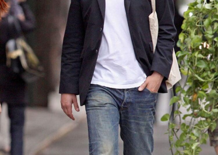 A man wearing a black fedora, sunglasses, black blazer, white t-shirt, blue jeans, and red sneakers walks on a city sidewalk with one hand in his pocket and a tote bag over his shoulder.