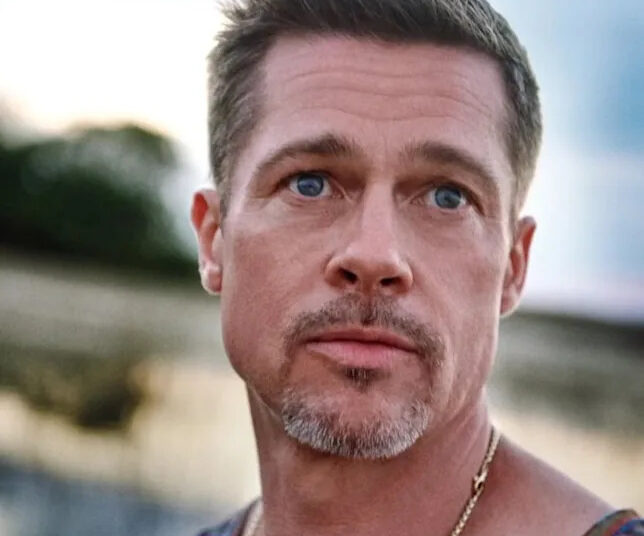 A man with short brown hair, blue eyes, and light facial hair gazes slightly to the side outdoors. He wears a sleeveless shirt and a gold chain necklace, with a blurred natural background behind him.
