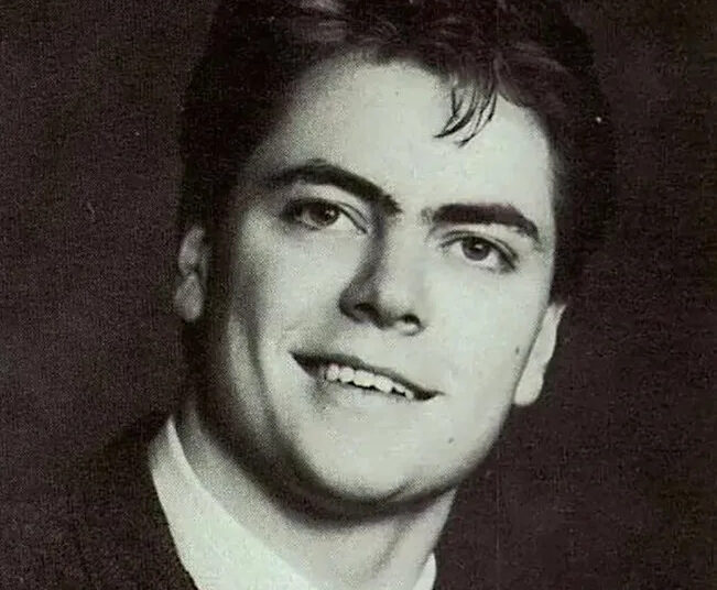 A young man in a black-and-white portrait, wearing a suit and tie. He has dark hair, combed back, and is smiling slightly while looking just off camera. The background is plain and dark.
