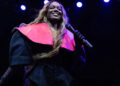 Azealia Banks on stage with a microphone, wearing a black outfit with a bright pink collar, under blue stage lights.