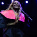 Azealia Banks on stage with a microphone, wearing a black outfit with a bright pink collar, under blue stage lights.