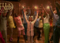 A group of people in colorful outfits are joyfully raising their hands on a stage. A child is singing into a microphone.