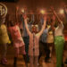 A group of people in colorful outfits are joyfully raising their hands on a stage. A child is singing into a microphone.