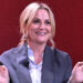Amy Poehler with blonde hair is smiling, wearing a gray blazer and white shirt, gesturing with her hands against a red background.