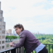 Person leaning on balcony railing with cityscape in background.
