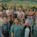 The choir is standing outside and looking at the camera, many toasting the picture with a pint. There are men and women with Jeremy Clarkson in the middle.