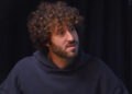 Lil Dicky with curly hair and a beard wearing a dark hoodie, sitting in front of a dark background.