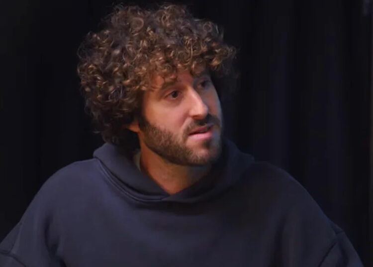 Lil Dicky with curly hair and a beard wearing a dark hoodie, sitting in front of a dark background.