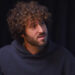 Lil Dicky with curly hair and a beard wearing a dark hoodie, sitting in front of a dark background.