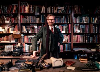 Mark Gatiss, as Gabriel Book, stands in front of a bookcase.