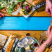 An overhead shot of a wooden table with an assortment of food arranged around the edges and two hands reaching across to exchange a slice of pizza.