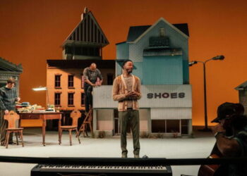 A Black man stands onstage; in the foreground a cellist plays with his back to the camera, and in the background two men work on a set that resembles a small town.
