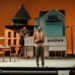 A Black man stands onstage; in the foreground a cellist plays with his back to the camera, and in the background two men work on a set that resembles a small town.