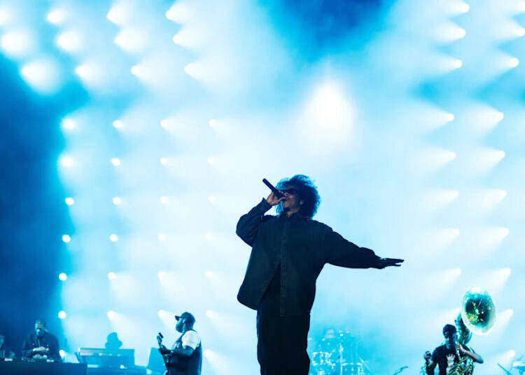 Jay-z on stage with bright blue lights in the background, musicians playing instruments on the side. This was at Roots Picnic 2026