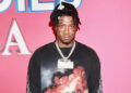 Blueface wearing a black shirt and a chain necklace stands in front of a pink background.