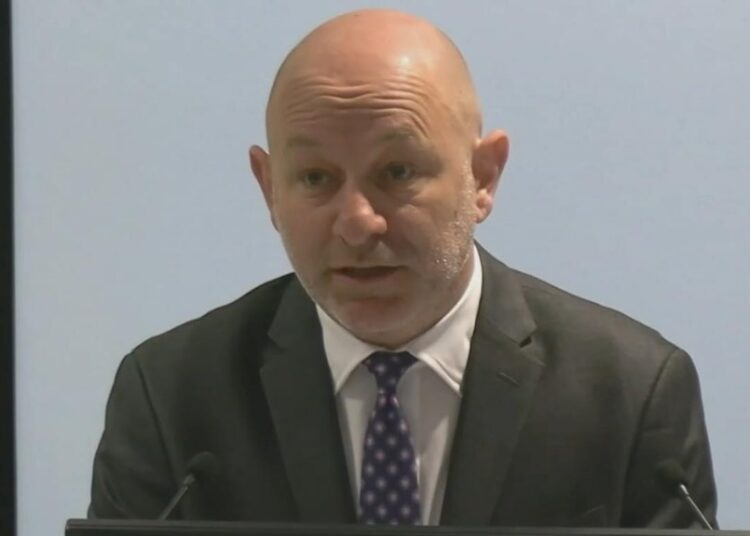 Man in suit speaks into microphone, grey background