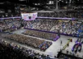 Wide view of a graduation ceremony in a packed indoor arena, with rows of graduates seated on the floor and a stage set up at the front.