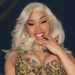 Cardi B with long blonde hair in a patterned outfit, smiling and posing with her hand near her mouth.