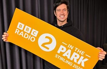 BBC Radio 2 presenter Vernon Kay holds a large, organge sign with branding for BBC Radio 2 In The Park for Stirling 2026.