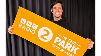 BBC Radio 2 presenter Vernon Kay holds a large, organge sign with branding for BBC Radio 2 In The Park for Stirling 2026.