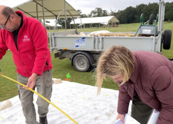 A man in a red fleece and woman wearing a padded coat, use a tape measure to transfer the garden plan onto the site, in front of a trailer filled with material for the build.