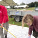 A man in a red fleece and woman wearing a padded coat, use a tape measure to transfer the garden plan onto the site, in front of a trailer filled with material for the build.
