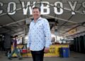 A man stands in front of a banner sign that reads "Cowboys" in silver capital letters hung from a tent with portapotties in the background.
