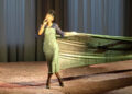 Lily Allen, with a large fabric wrapped around her like a dress, sings on stage. The backdrop is a curtain, and the stage floor is textured.