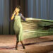 Lily Allen, with a large fabric wrapped around her like a dress, sings on stage. The backdrop is a curtain, and the stage floor is textured.