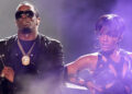 Sean "Diddy" Combs wearing sunglasses and a chain, performing on stage with Dawn Richard in a black outfit.