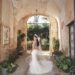 Bride in lace gown standing in a rustic archway, surrounded by greenery.