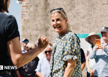 A picture of Sophie, Duchess of Edinburgh, meeting islanders. She is wearing a green and yellow short-sleeved blouse and she is wearing sunglasses on her head.