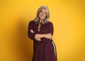 Sara Davies stood in front of a mustard yellow background, posing with her arms folded in front of her.
