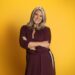 Sara Davies stood in front of a mustard yellow background, posing with her arms folded in front of her.