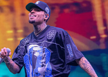 Chris Brown performing on stage, wearing a black graphic shirt and holding a microphone, with a colorful background.