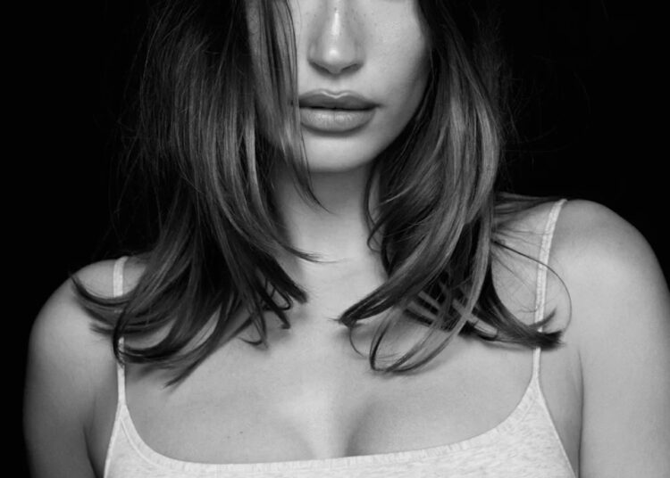 Black and white portrait of Hailey Bieber with long hair, wearing a light-colored tank top, looking directly at the camera.