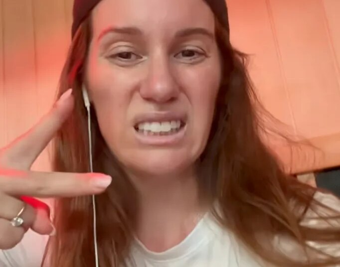 Comedian Hannah Berner making a sour face and wearing a black backward baseball cap, white t-shirt, and white wired earbuds.