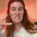 Comedian Hannah Berner making a sour face and wearing a black backward baseball cap, white t-shirt, and white wired earbuds.